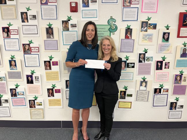 Lisa of Women's Empowerment wears a black jacket and pants and holds a check next to Pam Maxwell of US Bank wearing a blue dress in front of a wall covered with photos and stories of Women's Empowerment graduates