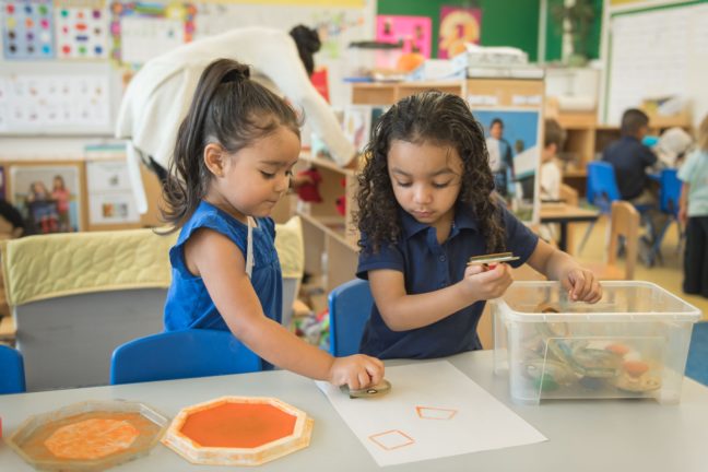 Two preschool girls play with stamps and an orange stamp pad to make shapes on a piece of white paper