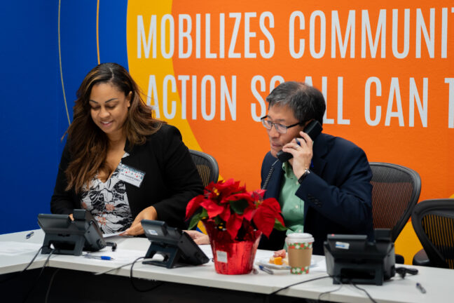 United Way president and CEO Dr. Dawnté Early and board chair Dr. Richard Pan make phone calls to donors under a new yellow blue and orange mural that reflects United Way’s refreshed brand.