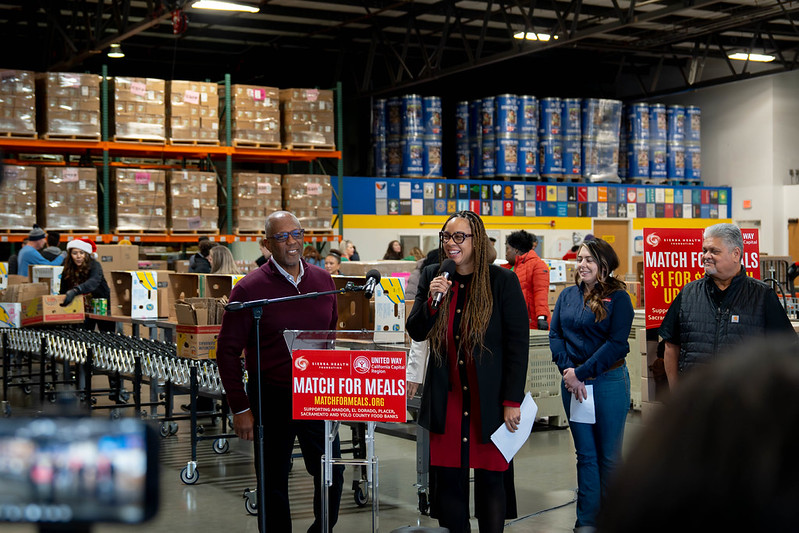 Dr. Early and Chet Hewitt speak at a podium in the warehouse at Sacramento Food Bank with volunteers packing food behind them